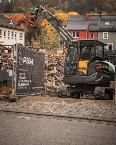 Volvo-Bagger ECR50 mit Sortiergreifer auf einer Abbruchstelle, im Hintergrund Bauzaun mit PSM-Werbung und Herbstb&auml;ume.