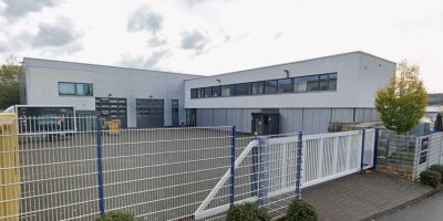 Modernes Firmengeb&auml;ude von PSM GmbH in Siegen mit grauer Fassade, Einfahrtstor und Metallzaun im Vordergrund.