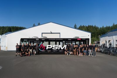 PSM Mitarbeiter vor einem PSM-Bus auf einem Parkplatz vor einem gro&szlig;en wei&szlig;en Industriegeb&auml;ude in Brachbach.