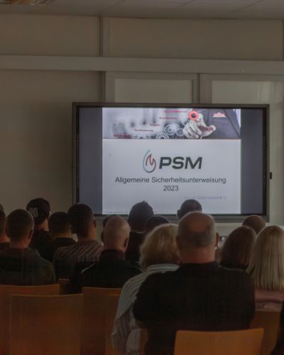 Zuschauer sitzen im Konferenzraum und schauen auf einen Bildschirm mit der Pr&auml;sentation &bdquo;Allgemeine Sicherheitsunterweisung 2023&ldquo;.