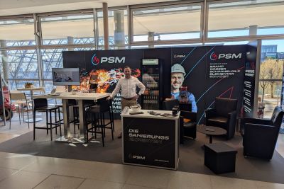 PSM-Messestand mit Bannerw&auml;nden, Stehtischen und Sitzbereich in einer Halle mit gro&szlig;en Fensterfronten.