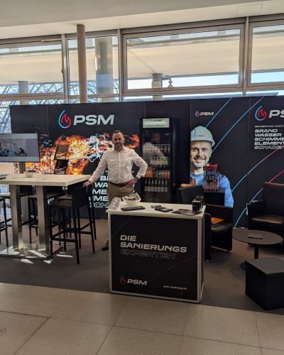 PSM-Messestand mit Bannerw&auml;nden, Stehtischen und Sitzbereich in einer Halle mit gro&szlig;en Fensterfronten.