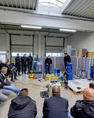 Gruppe von PSM-Mitarbeitern sitzt und steht in einer Werkhalle w&auml;hrend einer Schulung oder Pr&auml;sentation zu technischem Equipment.