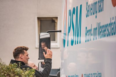Ein Mann in Arbeitskleidung steht neben einem gro&szlig;en wei&szlig;en PSM-LKW und blickt in den Seitenspiegel.