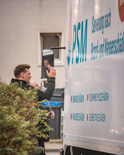Ein Mann in Arbeitskleidung steht neben einem gro&szlig;en wei&szlig;en PSM-LKW und blickt in den Seitenspiegel.