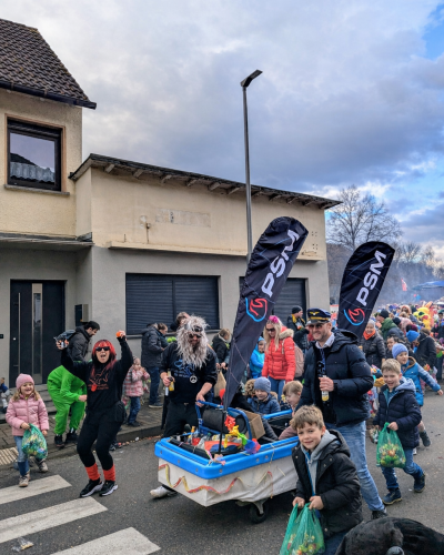 Menschen bei einem Stra&szlig;enumzug mit PSM-Werbefahnen, Kindern und einem geschm&uuml;ckten Bollerwagen vor einem Wohnhaus.