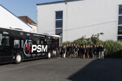 Schwarzer PSM-Bus vor einem Geb&auml;ude, daneben eine Gruppe von PSM Mitarbeitern in schwarzer Kleidung.