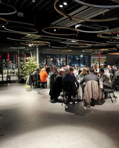 G&auml;ste sitzen an langen Tischen in einem modernen Veranstaltungsraum mit runden Deckenleuchten und Bar im Hintergrund.